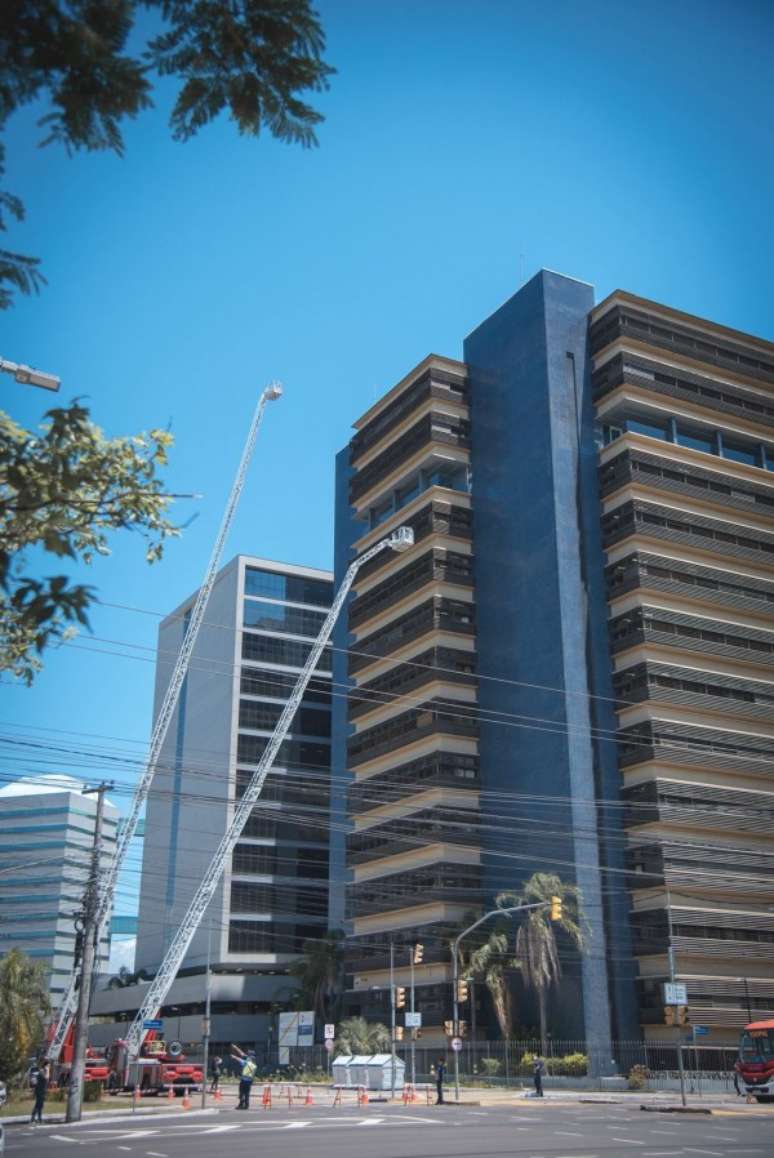 A foto duas autoescadas mecânicas de caminhões do Corpo de Bombeiros, estendidas em direção aos andares superiores de um edifício. Uma das escadas alcança quase o topo do prédio, enquanto a outra está posicionada um pouco mais abaixo.