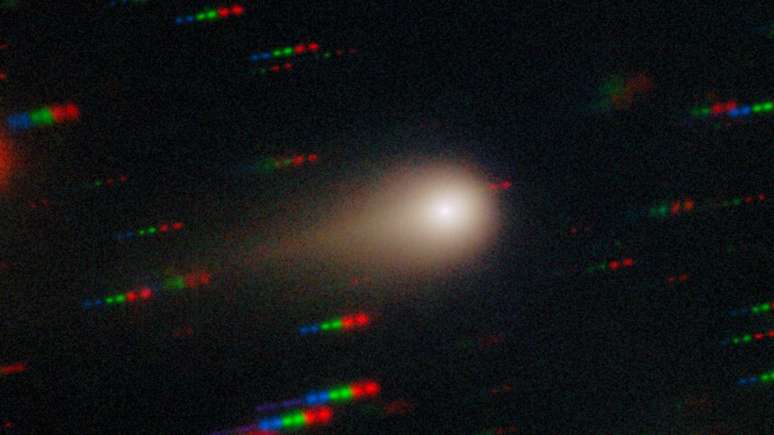 O cometa 3I/Atlas exibido na forma de núcleo brilhante, rodeado por uma coroa nebulosa e uma cauda à esquerda. Ao fundo, filas de pontos azuis, verdes e vermelhos, representando estrelas que se movem em relação ao cometa.