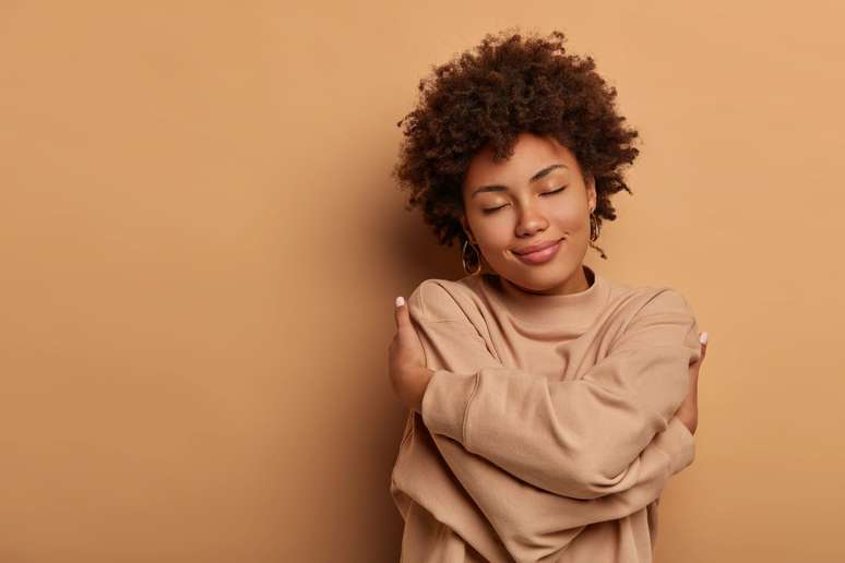 Love yourself, self acceptance. Gentle lovely Afro American woman crosses hands and embraces own body, tilts head and closes eyes, wears comforable brown sweater, models indoor. I love myself