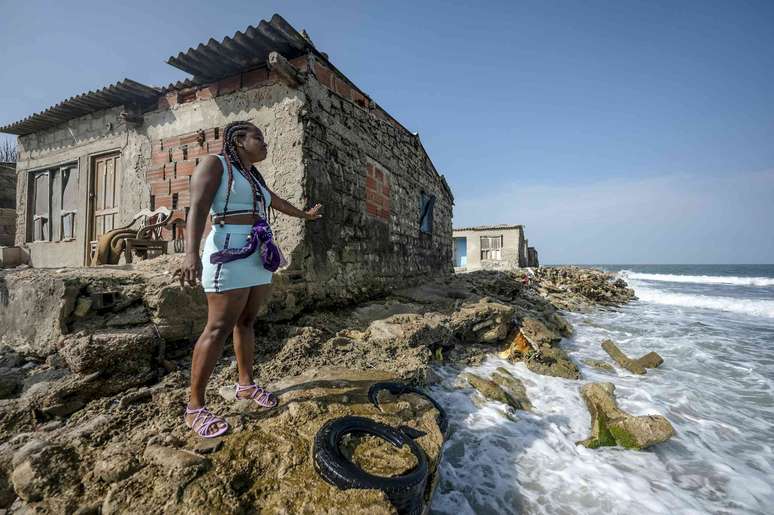Uma mulher em frente a uma casa antiga, mostrando onde o aumento do nível do mar erodiu a costa quase até a fundação da casa.