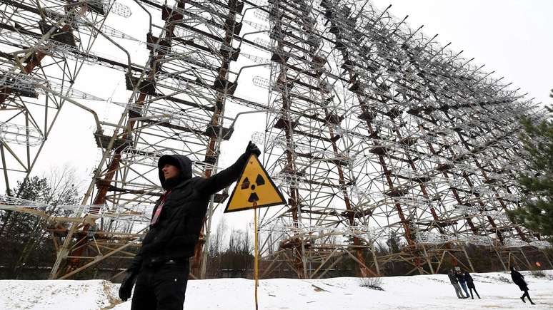 Pessoa de pé em frente a uma enorme antena de radar, ao lado de uma placa com o símbolo de radiação