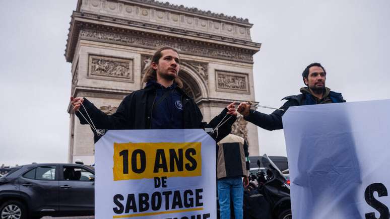 Ativista segura faixa com os dizeres “10 anos de sabotagem climática” durante ação do Greenpeace em frente ao Arco do Triunfo, em Paris, em 12 de dezembro de 2025, marcando o 10º aniversário da assinatura do Acordo Climático de Paris.