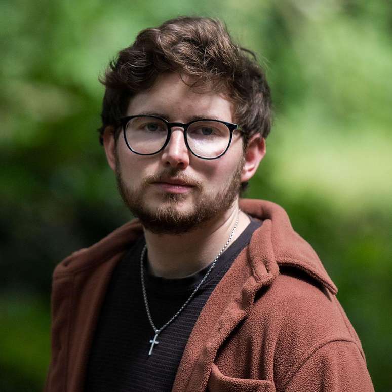 Homem de casaco marrom com camiseta preta e um crucifixo, em p&eacute;, usando &oacute;culos em frente a um arbusto