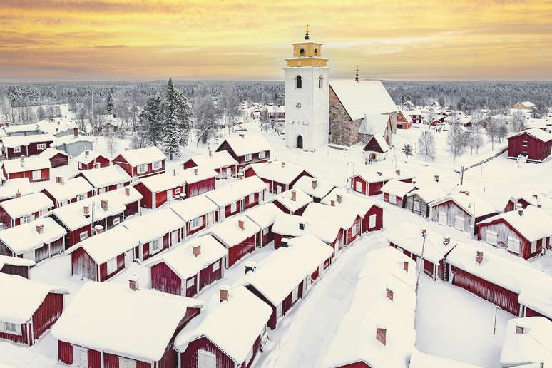 Vista a&eacute;rea dos telhados nevados de casas vermelhas e da torre sineira sob um c&eacute;u flamejante ao amanhecer, cidade da igreja de Gammelstad, Lule&aring;, Su&eacute;cia