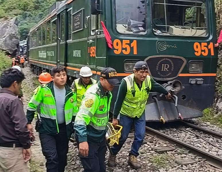 Membros das equipes de resgate, vestindo coletes amarelos, carregam uma pessoa ferida em uma maca ao lado de um trem.