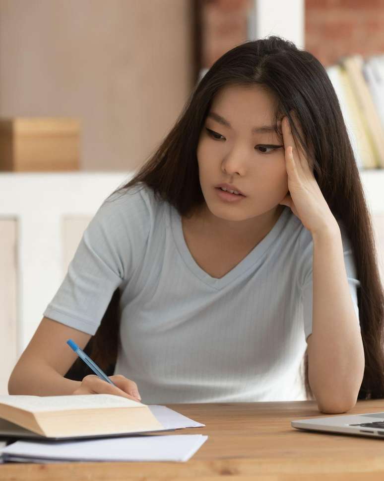 Mulher asi&aacute;tica jovem sentada na cadeira mexendo em v&aacute;rios livros na mesa.