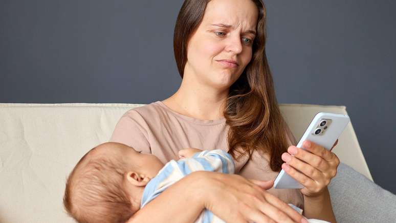 Mulher com express&atilde;o triste, sentada em um sof&aacute; bege, segurando um beb&ecirc; no colo com roupa listrada enquanto usa o telefone celular.
