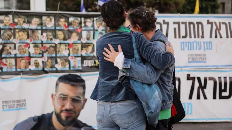 Mulheres israelenses se abra&ccedil;am diante de um banner com fotos de ref&eacute;ns do grupo Hamas, entre elas, a do policial israelense Ran Gvili. Tel Aviv, 26 de janeiro de 2026.