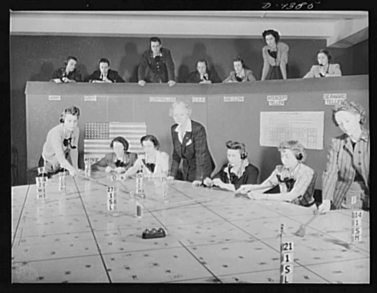 Fotografia em preto e branco de mulheres em torno de uma mesa quadriculada, uma pessoa empurrando pe&ccedil;as pela superf&iacute;cie enquanto outras observam em uma varanda