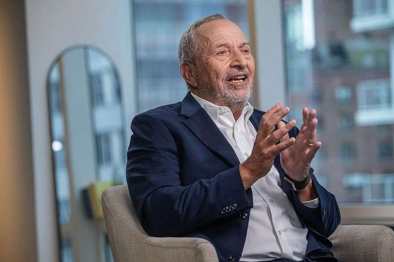 Larry Summers est&aacute; sentado em uma poltrona cinza, em um apartamento. Atr&aacute;s dele h&aacute; um espelho. Ele usa um terno azul e uma camisa branca e gesticula com as m&atilde;os enquanto fala. 