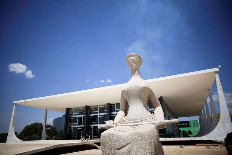 Est&aacute;tua de pedra da Justi&ccedil;a em primeiro plano, sentada e segurando uma espada, em frente ao pr&eacute;dio do Supremo Tribunal Federal, em Bras&iacute;lia, sob c&eacute;u azul intenso.
