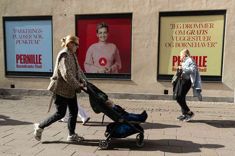 Uma mulher empurra um carrinho com um beb&ecirc; passando por um cartaz eleitoral, enquanto outra mulher cruza seu caminho carregando objetos de forma desajeitada.