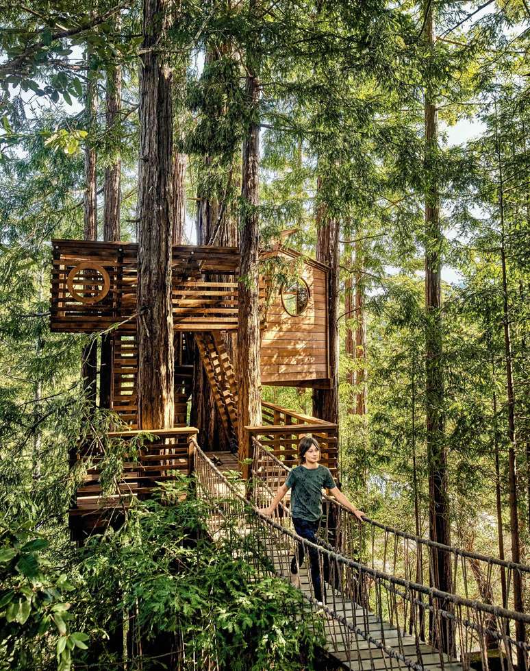 Uma casa na &aacute;rvore de madeira com dois andares, conectada a uma ponte suspensa sobre a qual uma mulher caminha.