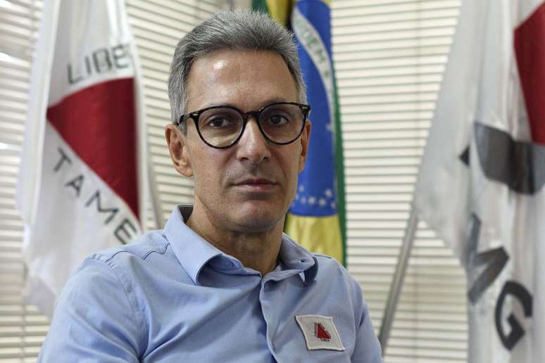 Romeu Zema &eacute; um homem branco e usa &oacute;culos de grau com arma&ccedil;&atilde;o preta. Ele est&aacute; com uma camisa azul clara que tem uma bandeira de Minas Gerais. 