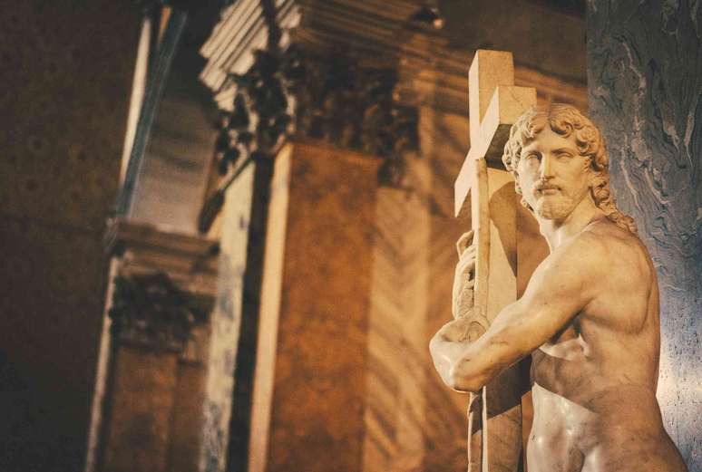 est&aacute;tua de pedra de homem segurando uma grande cruz, em ambiente de igreja