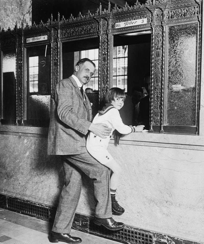 Amadeo Giannini carrega o ator infantil Jackie Coogan (1914-1984) &mdash; do filme de Chaplin O Garoto (1921) &mdash; para alcan&ccedil;ar o guich&ecirc; do caixa do banco