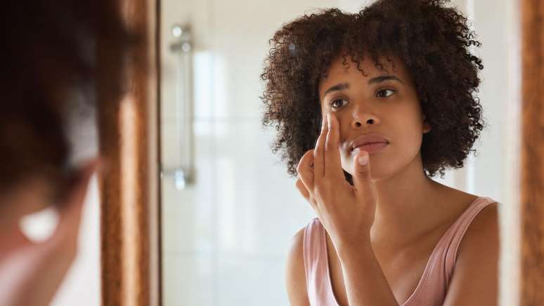 ముదురు బొచ్చు గల యువతి అద్దంలో తనను తాను చూసుకుంటూ, తన కళ్ల చుట్టూ ఉన్న చర్మాన్ని దిగులుగా చూస్తూ