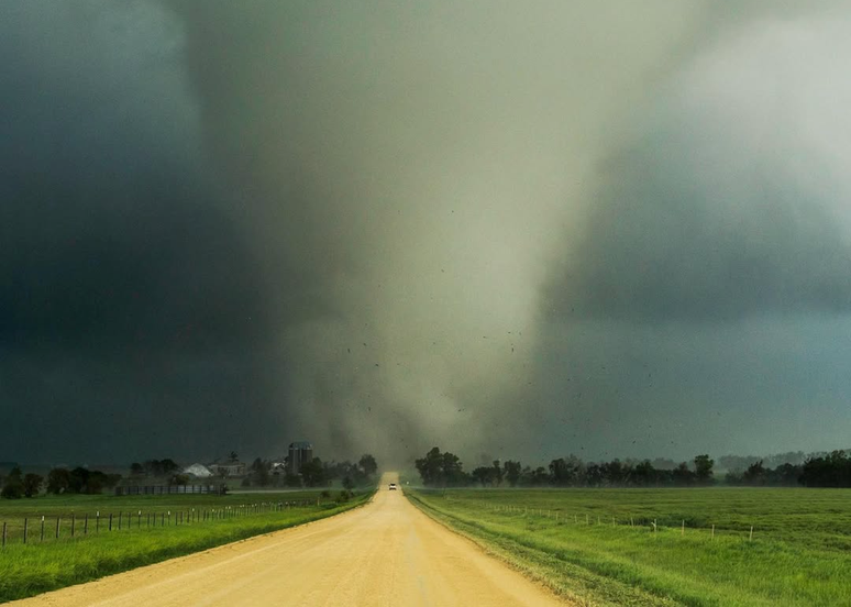 Um ve&iacute;culo desce por uma estrada rural, afastando-se de um grande tornado.