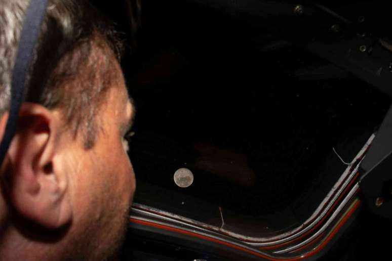 Um astronauta olhando pela janela da espa&ccedil;onave Orion, de onde a lua cheia &eacute; vis&iacute;vel no espa&ccedil;o.