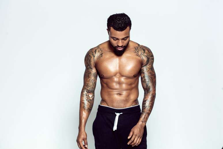 Homem com os bra&ccedil;os tatuados, sem camisa, olhando para baixo