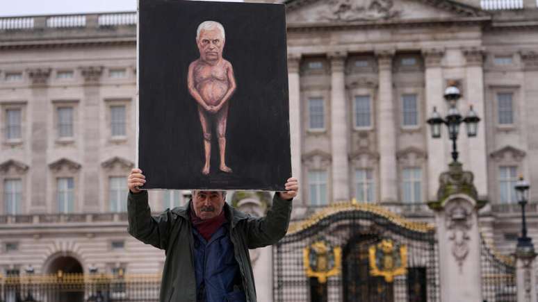 O artista Kaya Mar posa com a mais recente edi&ccedil;&atilde;o de suas pinturas para a m&iacute;dia em frente ao Pal&aacute;cio de Buckingham, em Londres, na sexta-feira, 20 de fevereiro de 2026, ap&oacute;s Andrew Mountbatten-Windsor ter sido preso e detido por horas pela pol&iacute;cia brit&acirc;nica sob suspeita de m&aacute; conduta no exerc&iacute;cio de cargo p&uacute;blico relacionada a suas liga&ccedil;&otilde;es com Jeffrey Epstein.