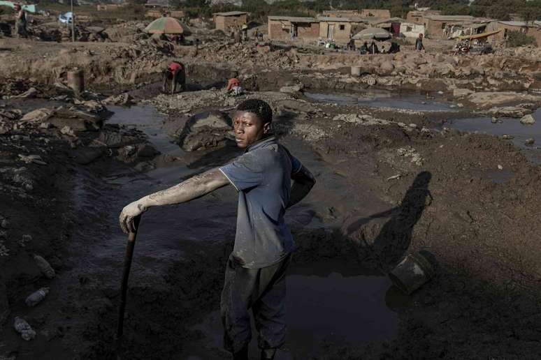Um homem com lama seca nos bra&ccedil;os nus est&aacute; perto de uma mina cheia de &aacute;gua onde uma crian&ccedil;a e uma mulher procuram minerais.