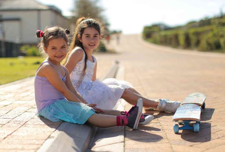 Duas crian&ccedil;as vestidas com vestidos sentadas na cal&ccedil;ada de um bairro, com skates &agrave; sua frente