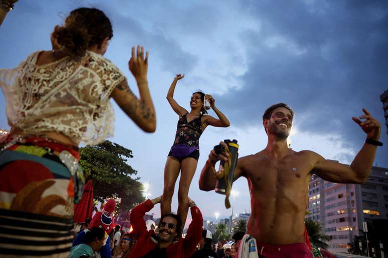 Pessoas dan&ccedil;am com os bra&ccedil;os para a cima na praia. Um homem est&aacute; sem camisa. Outro carrega uma mulher nos ombros. 