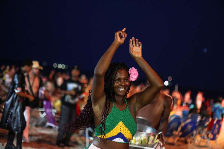 Mulher negra com blusa nas cores do Brasil. Ela levanta os bra&ccedil;os e sorri, com multid&atilde;o ao fundo