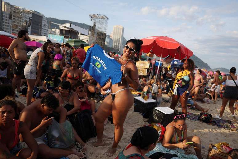 Mulher de biqu&iacute;ni segura camiseta com o nome Shakira e uma bandeira do Brasil. Est&aacute; na praia, de dia. 