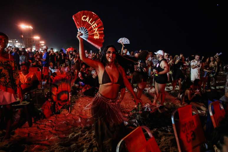 Mulher segura leque com a palavra caliente. Ela est&aacute; na praia, &agrave; noite. Muita gente atr&aacute;s. 