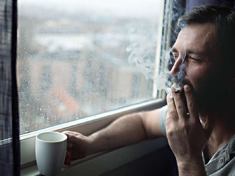 Homem tomando café e fumando um cigarro ao lado de uma janela.