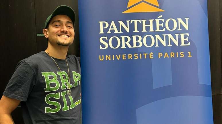 O estudante brasileiro Caio D&eacute;rio ao lado de uma placa com o nome da universidade onde estuda, Panth&eacute;on Sorbonne, que subiu as taxas de matr&iacute;cula em dezembro de 2025.