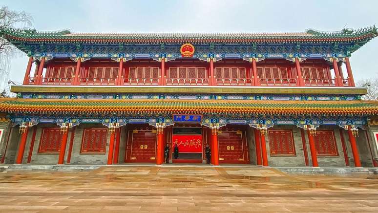 Um edif&iacute;cio tradicional chin&ecirc;s, com pilares vermelhos e grandes port&otilde;es vermelhos ao centro; v&aacute;rios guardas permanecem de p&eacute; na entrada.
