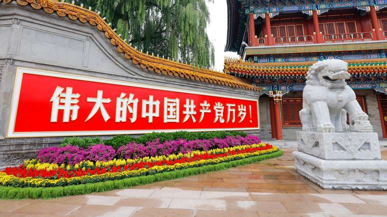 Um letreiro vermelho com caracteres chineses brancos est&aacute; pendurado em um muro de pedra, com um colorido canteiro de flores abaixo; um le&atilde;o de pedra branca est&aacute; em cima de um pedestal ao lado, enquanto um edif&iacute;cio ornamentado aparece ao fundo.