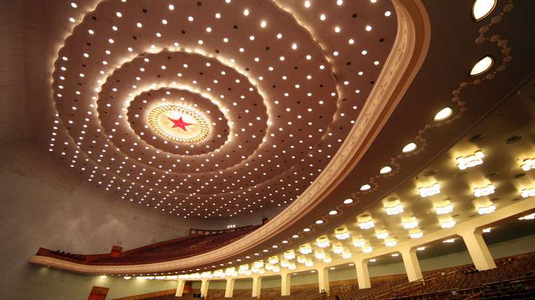 Imagem do interior de um audit&oacute;rio mostra um teto repleto de luzes brancas e uma estrela vermelha com um c&iacute;rculo iluminado no centro. Tamb&eacute;m &eacute; poss&iacute;vel ver fileiras de assentos.

