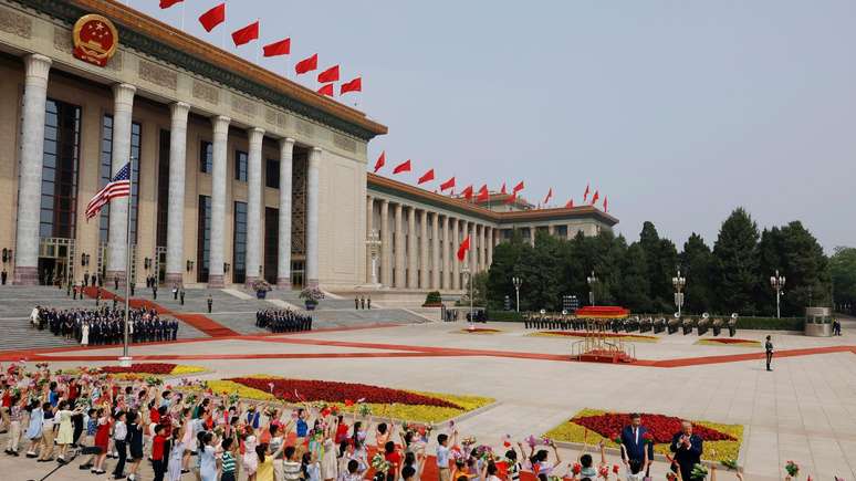 Vista de uma pra&ccedil;a pavimentada, com bandeiras chinesas e americanas hasteadas, e um longo edif&iacute;cio com colunas &agrave; esquerda, ostentando bandeiras chinesas no teto. Em primeiro plano, os presidentes Xi e Trump acenam para a multid&atilde;o.
