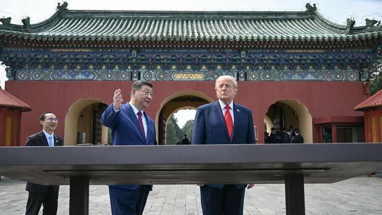 Os presidentes Xi e Trump se encontram no p&aacute;tio de um complexo templ&aacute;rio, atr&aacute;s de uma grande exposi&ccedil;&atilde;o, com Xi levantando a m&atilde;o para mostrar algo a Trump.
