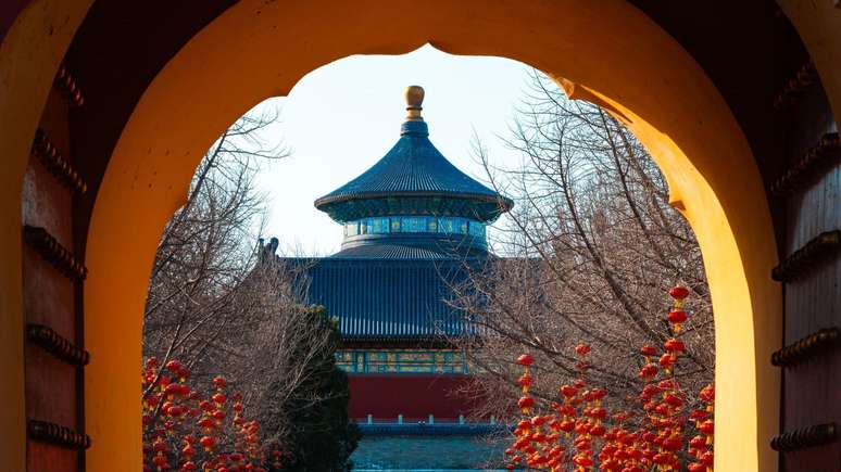 Uma vista atrav&eacute;s de um portal hist&oacute;rico de onde se v&ecirc; edif&iacute;cios de um templo em tons de azul, vermelho e dourado, com numerosas lanternas chinesas penduradas nas &aacute;rvores sem folhas em primeiro plano.
