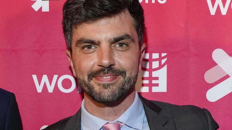 Will Adolphy, um homem com barba e cabelo curto, vestido com terno, camisa azul e gravata rosa. Ele sorri olhando diretamente para a c&acirc;mera.