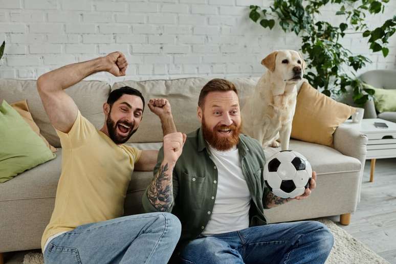 Dois homens, sentados no ch&atilde;o e assistindo futebol, cachorro sentado no sof&aacute; ao lado