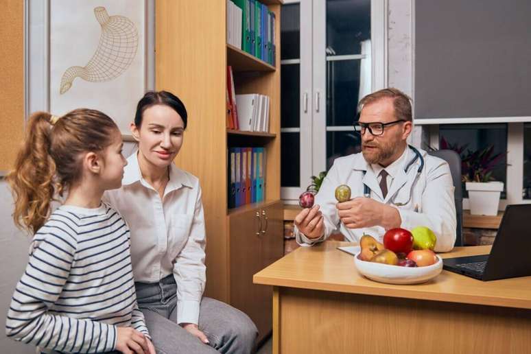 M&eacute;dico de jaleco e &oacute;culos em seu consult&oacute;rio mostra duas frutas pequenas para uma menina e sua m&atilde;e, que est&atilde;o sentadas &agrave; sua frente ouvindo atentamente. Sobre a mesa, h&aacute; uma fruteira cheia de ma&ccedil;&atilde;s e peras.