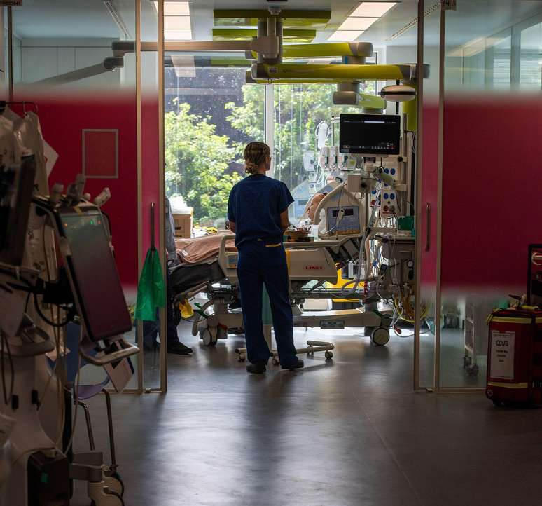 Hollie, de 29 anos, est&aacute; deitada sobre travesseiros em sua cama de hospital na UTI do King's College Hospital; ela veste um avental hospitalar com tubos conectados ao nariz, enquanto sua m&atilde;e est&aacute; sentada ao lado da cama (fora do enquadramento) e uma enfermeira a examina.