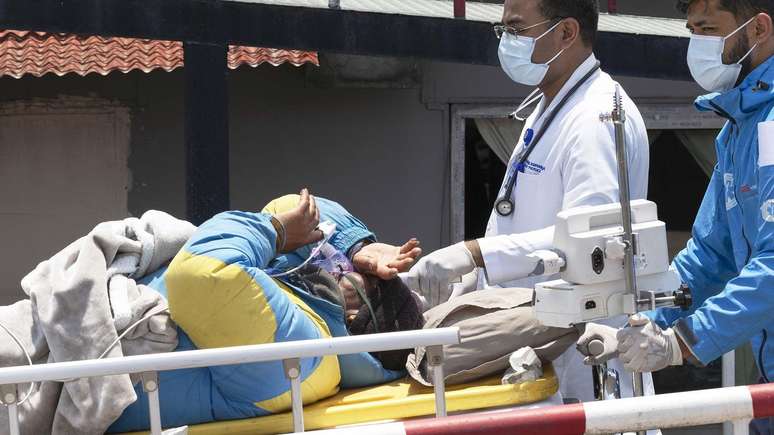 Dawa Sherpa levado em uma maca acompanhado por dois m&eacute;dicos. Ele veste uma jaqueta de montanhismo amarela e azul 