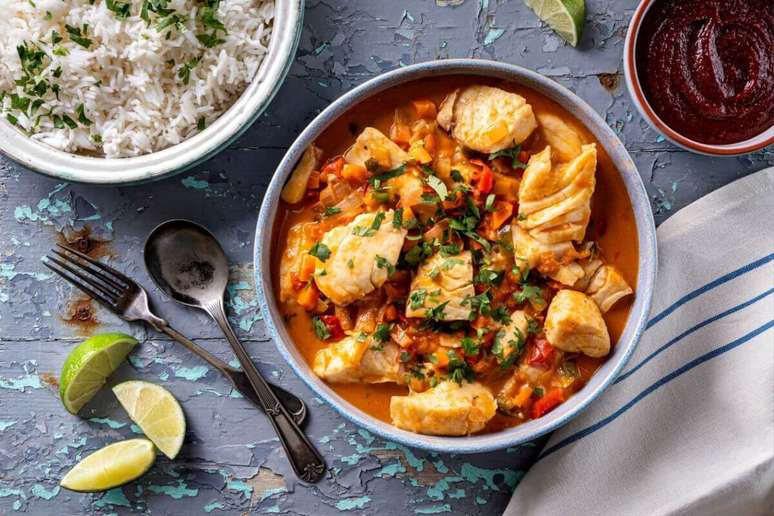 Uma Moqueca de Peixe vibrante, com peda&ccedil;os suculentos de peixe branco em um molho alaranjado de tomates e piment&otilde;es, finalizada com coentro fresco. Uma tigela de arroz branco soltinho e um pequeno bowl com molho de pimenta (ou pir&atilde;o) bem avermelhado.
