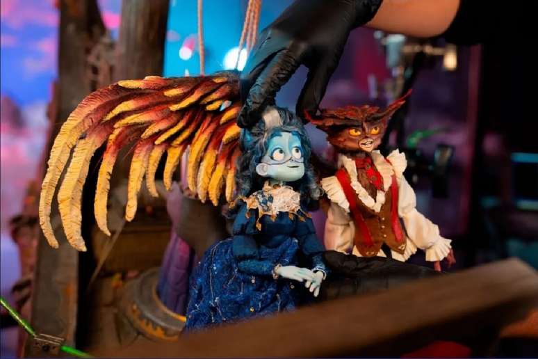 astidores de uma produ&ccedil;&atilde;o em stop-motion. Uma m&atilde;o usando luva cir&uacute;rgica preta ajusta delicadamente a cabe&ccedil;a de uma boneca com pele azul claro, cabelos escuros e vestido azul imperial de &eacute;poca. Ao lado dela est&aacute; outro boneco com fei&ccedil;&otilde;es de coruja vestindo colete marrom. O cen&aacute;rio de fundo &eacute; desfocado.