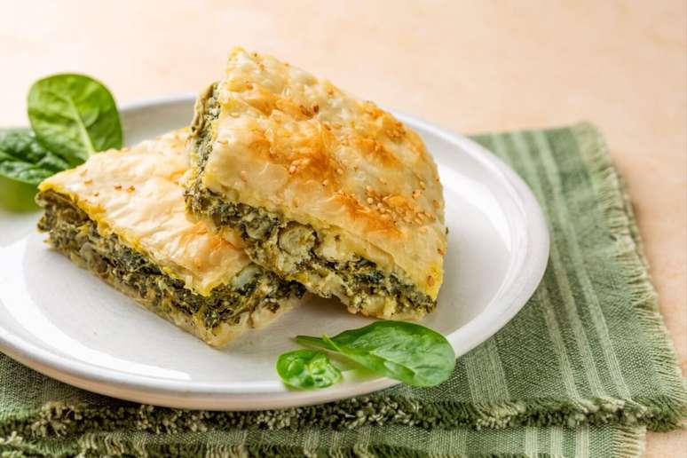 A imagem mostra dois peda&ccedil;os de uma torta salgada assada, com massa fina e dourada, coberta por sementes, possivelmente gergelim. O recheio parece ser cremoso e verde, lembrando espinafre