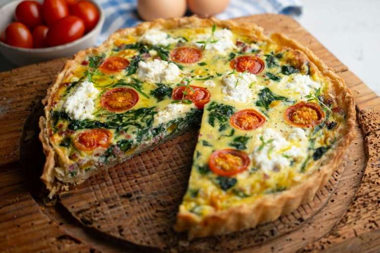 A imagem mostra uma quiche assada com apar&ecirc;ncia caseira e recheio colorido e cremoso. A massa parece crocante e dourada, enquanto o recheio leva ingredientes que lembram folhas verdes, possivelmente espinafre, tomates-cereja cortados ao meio e peda&ccedil;os de queijo branco espalhados por cima.