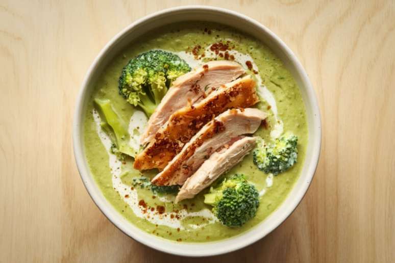 A imagem mostra uma tigela com um creme verde espesso, aparentemente feito &agrave; base de vegetais, acompanhado por peda&ccedil;os de frango fatiado e floretes de br&oacute;colis.