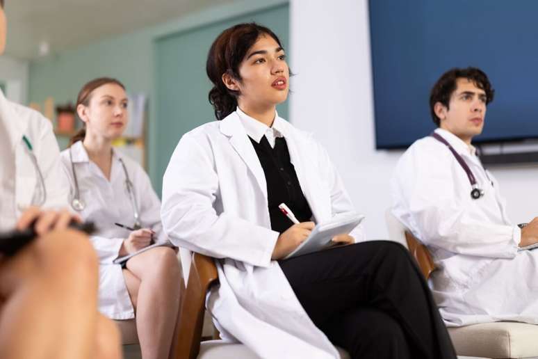 Em uma sala de aula ou audit&oacute;rio, uma jovem estudante de medicina com cabelos escuros presos est&aacute; sentada, olhando atentamente para frente, como se assistisse a uma palestra. Ela veste jaleco branco sobre uma blusa preta e camisa social, e segura um bloco de notas e uma caneta. Ao seu redor, outros estudantes de jaleco tamb&eacute;m prestam aten&ccedil;&atilde;o na aula, com uma tela de TV preta vis&iacute;vel ao fundo.